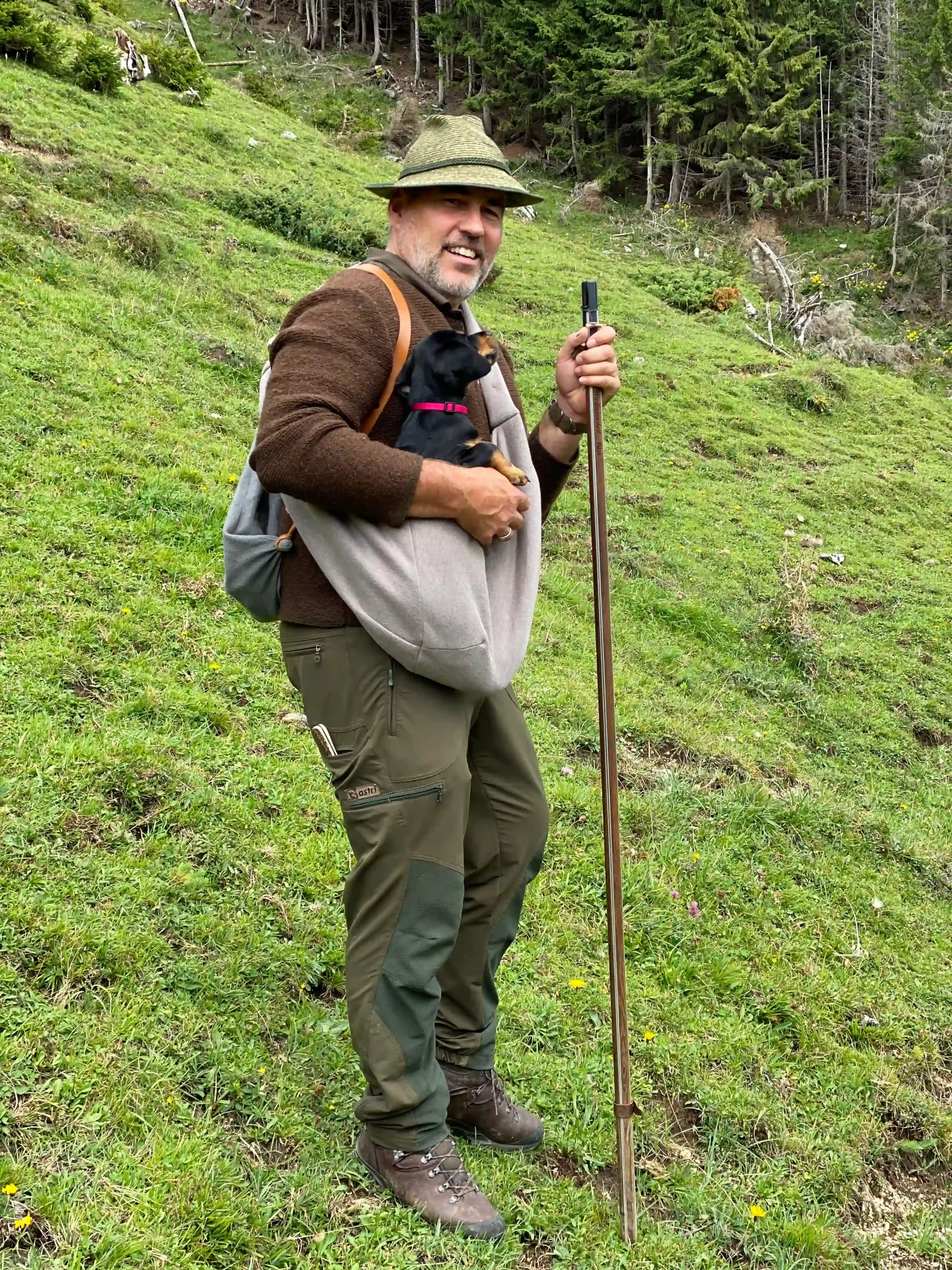 Harald Huber steht in alpiner Wiese mit Wanderstock und Tasche, Outdoor und Naturerlebnis